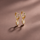 A pair of gold huggie hoop earrings with marquise-cut clear gemstones, displayed on a textured brown surface with soft shadows.