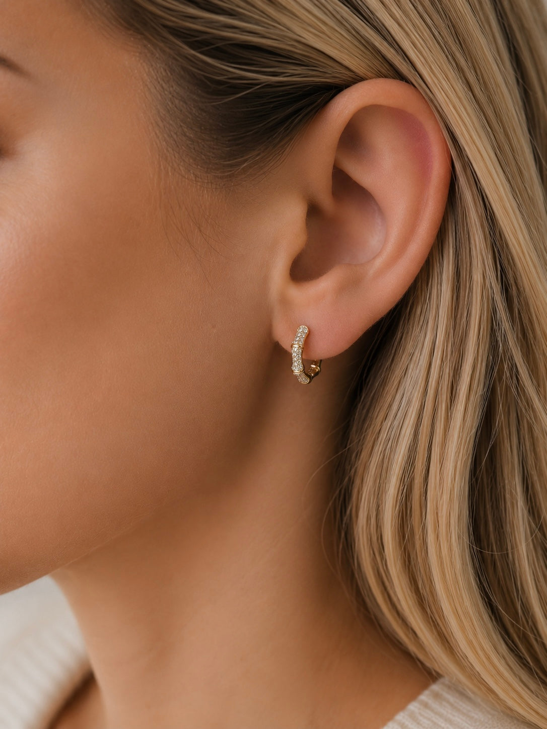 A close-up of a model's ear wearing a small gold huggie hoop earring adorned with tiny clear stones.