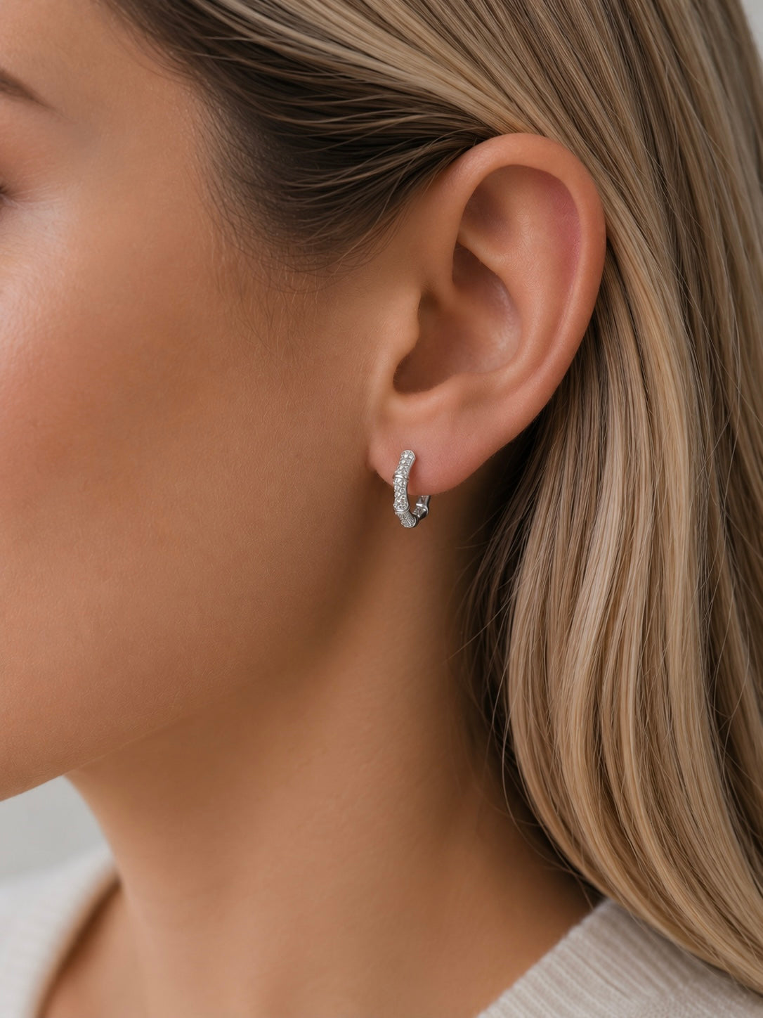 A close-up of a model's ear wearing a small silver huggie hoop earring set with clear stones.