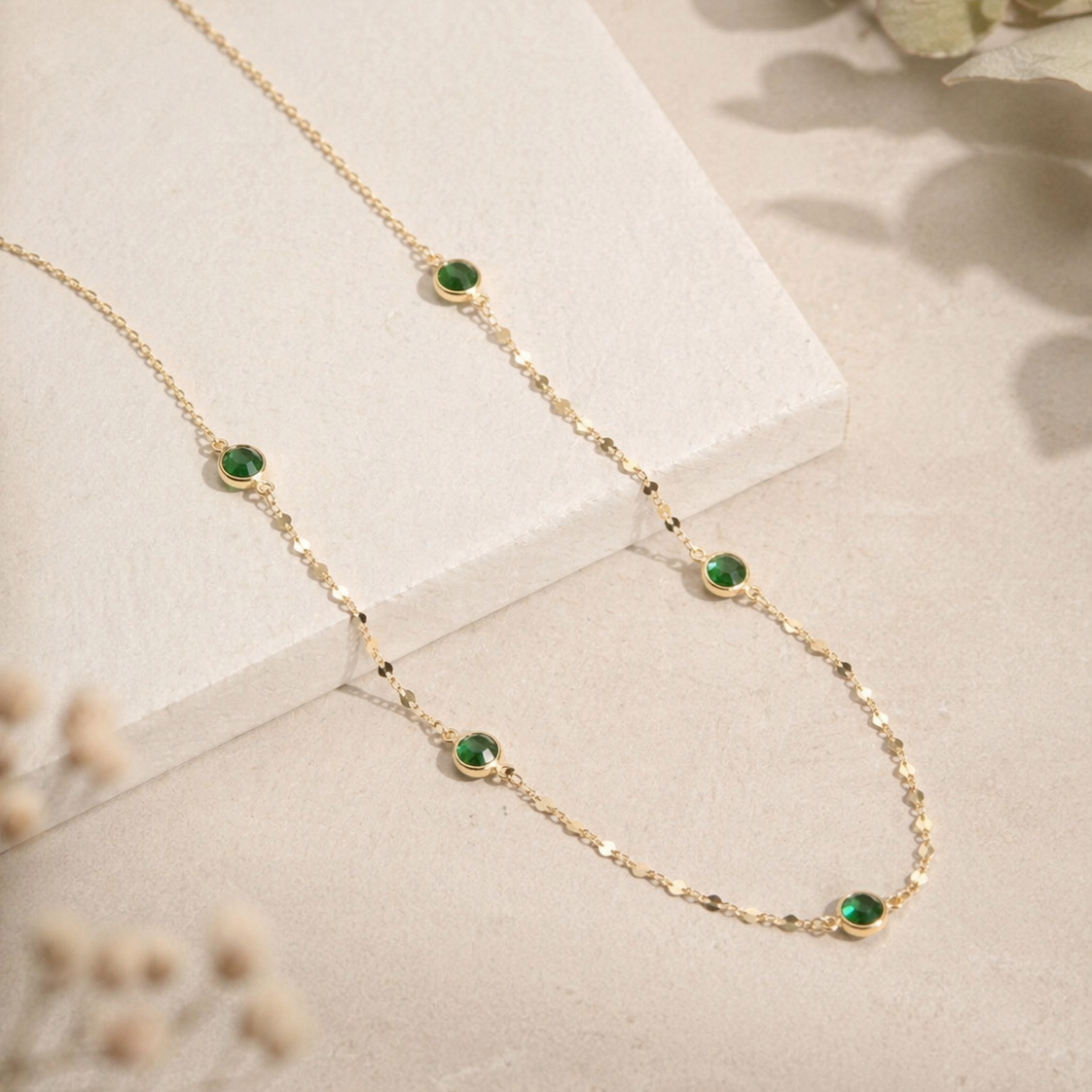 Gold necklace with green gemstones on a light surface with floral background